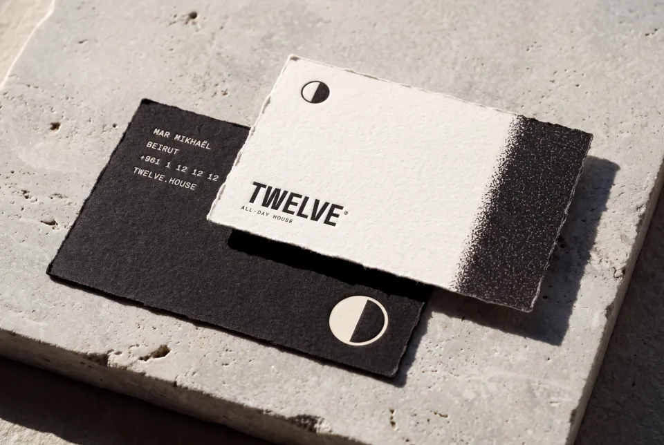 The TWELVE business cards on a stone slab in raking sun, the bone face with the mark and wordmark over the ink reverse