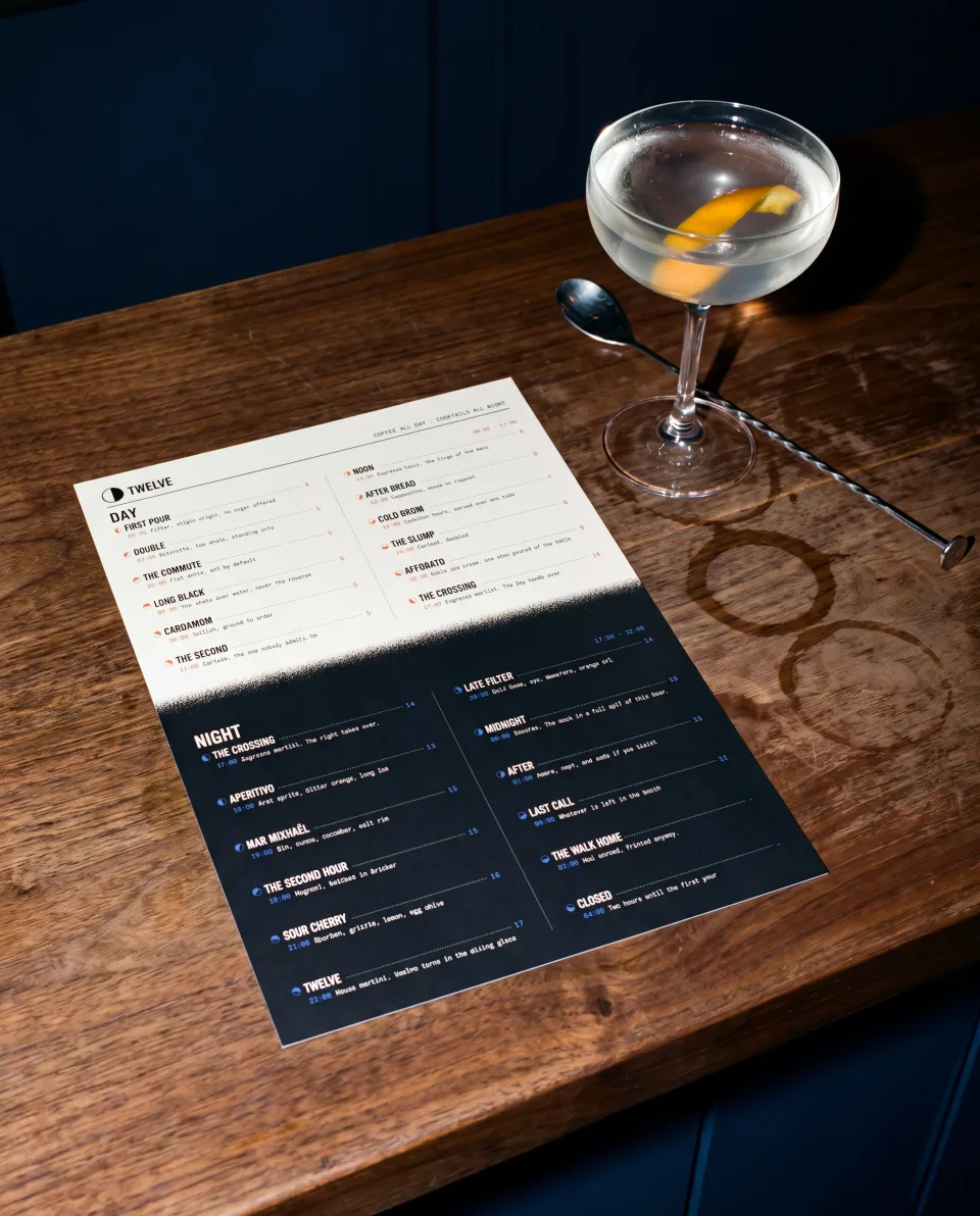 The same TWELVE all-day menu card on a walnut bar at night beside a coupe glass, caught by a direct flash