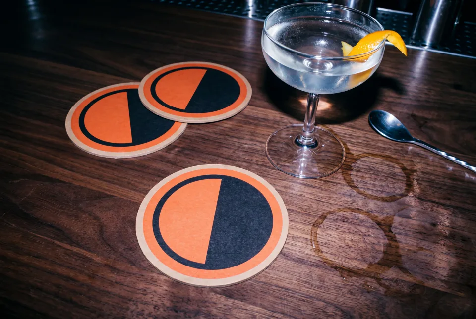 Three printed TWELVE coasters on a walnut bar beside a coupe glass, shot with direct flash late at night