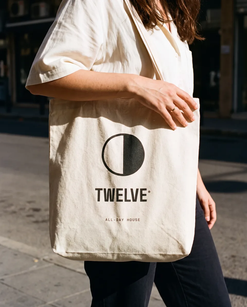 A natural canvas tote screen printed with the TWELVE mark and wordmark, carried on a Beirut street in daylight