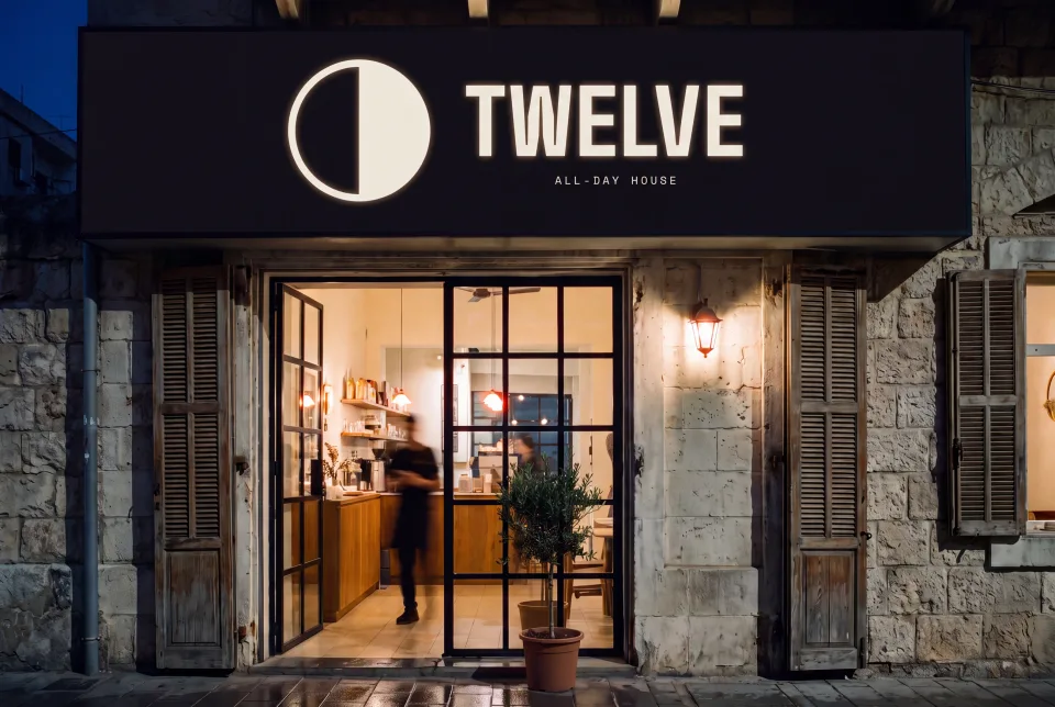 The same TWELVE shopfront at night, the sign panel now inverted to near-black with the mark and wordmark glowing bone out of it, over a wet street