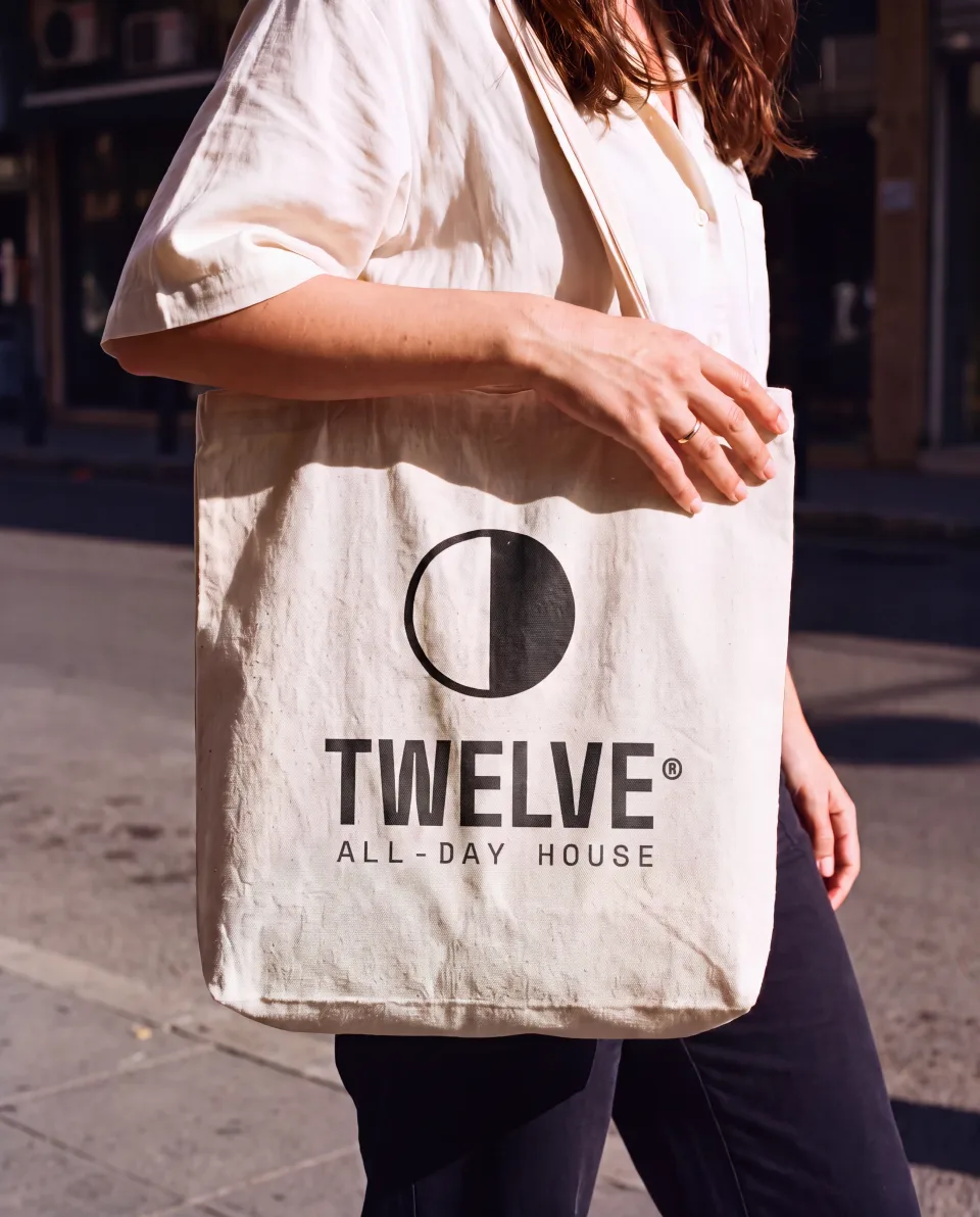 A natural canvas tote screen printed with the TWELVE mark and wordmark, carried on a Beirut street in daylight