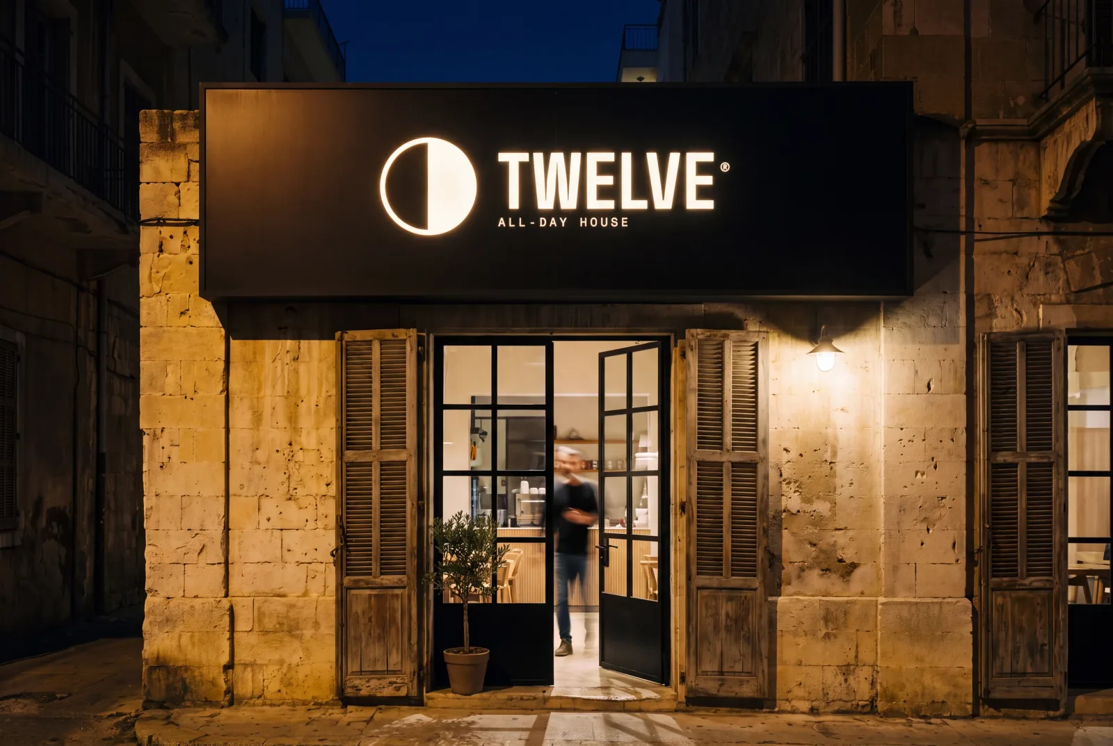 The same TWELVE shopfront at night, the sign now a glowing lightbox reading bone on black over a wet street