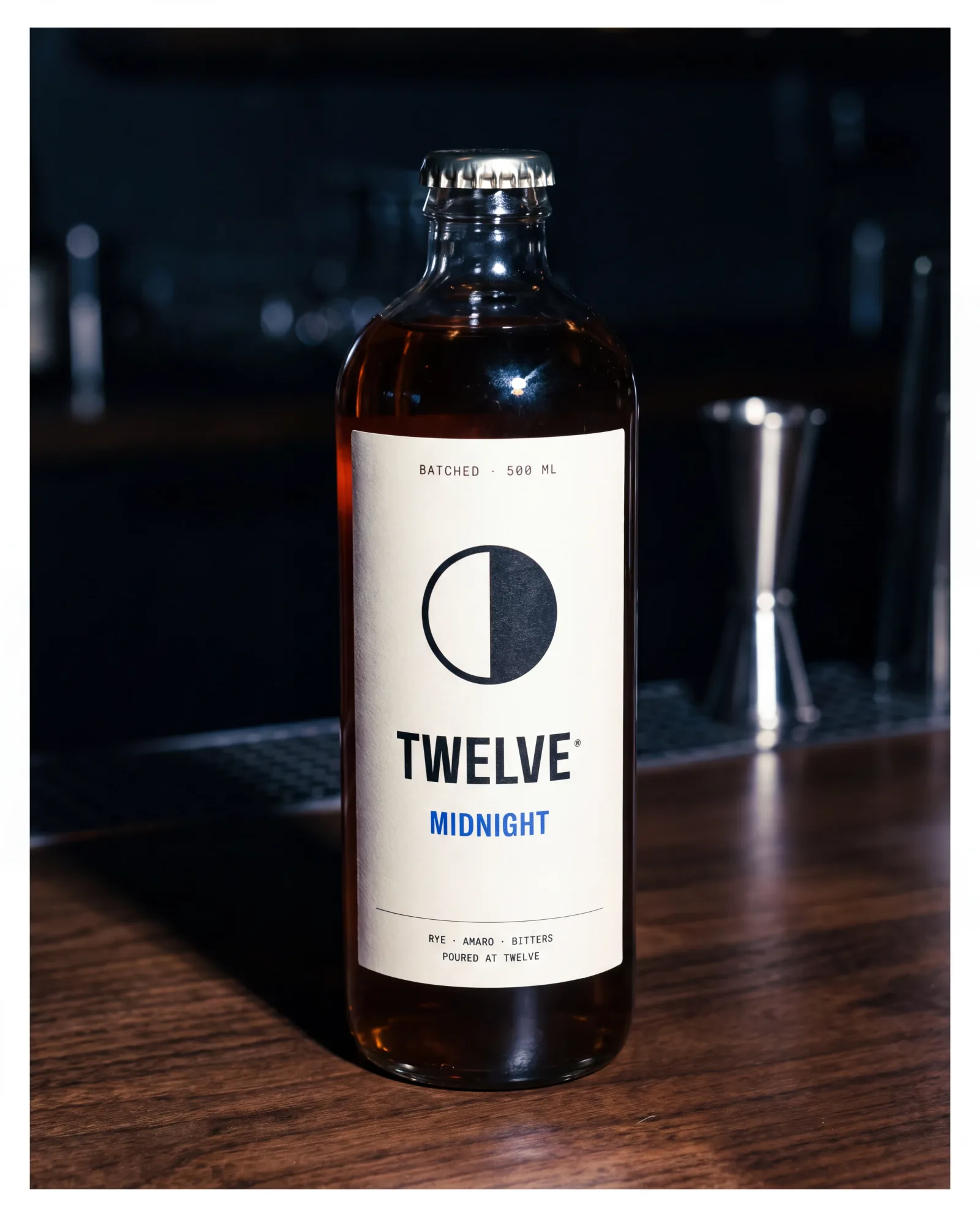 A TWELVE bottle of batched Midnight cocktail on a walnut bar, lit by direct flash against the dark room