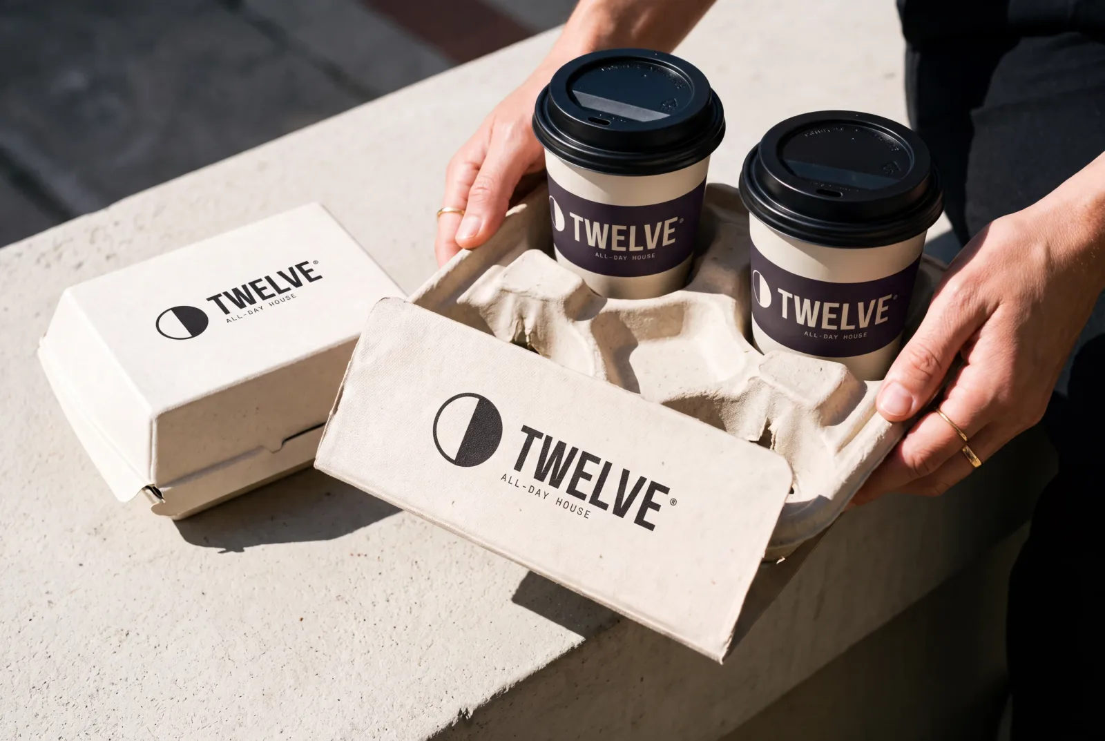 A pulp four-cup carrier holding two TWELVE takeaway cups, carried in two hands, with a printed takeaway box on the ledge beside it