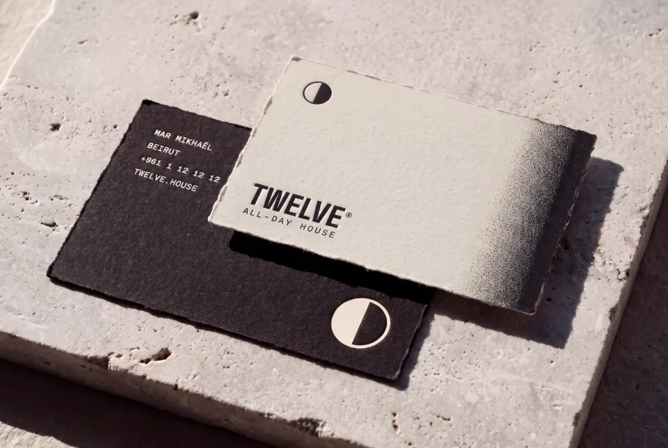The TWELVE business cards on a stone slab in raking sun, the bone face with the mark and wordmark over the ink reverse