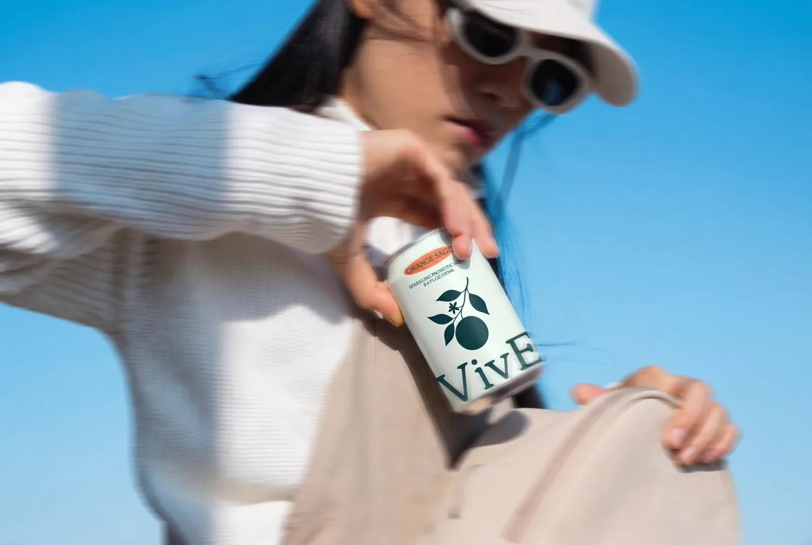 A woman in a white cap and sunglasses lifting a VivE Orange Sage can against a clear blue sky, the frame blurred by motion