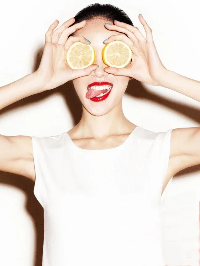 A woman holding two lemon slices over her eyes, tongue out, on white