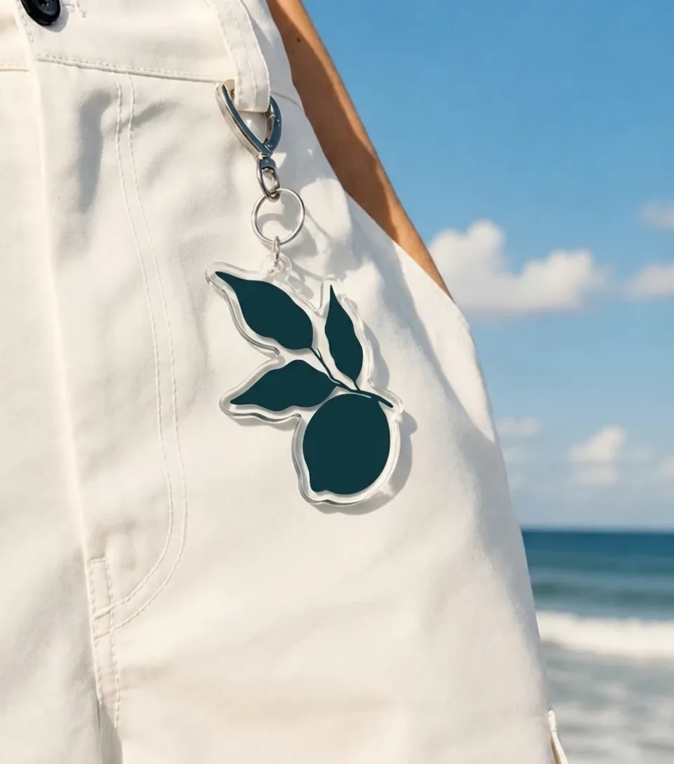 A clear acrylic VivE keychain carrying the lemon silhouette, clipped to white trousers with the sea behind