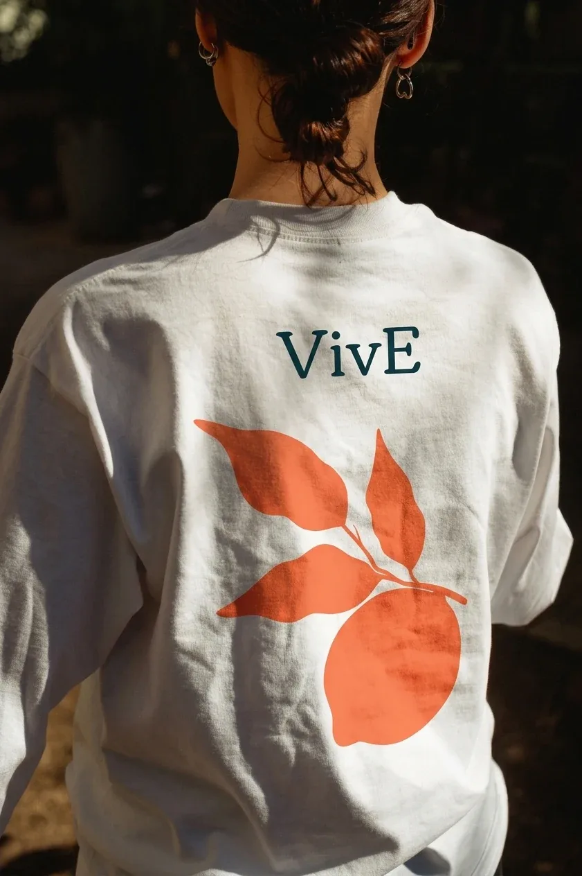 The back of a white VivE t-shirt in dappled shade, the wordmark in teal above a large orange lemon silhouette