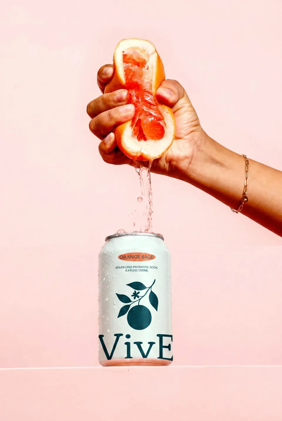 A hand squeezing half an orange over an open VivE Orange Sage can against a pink wall